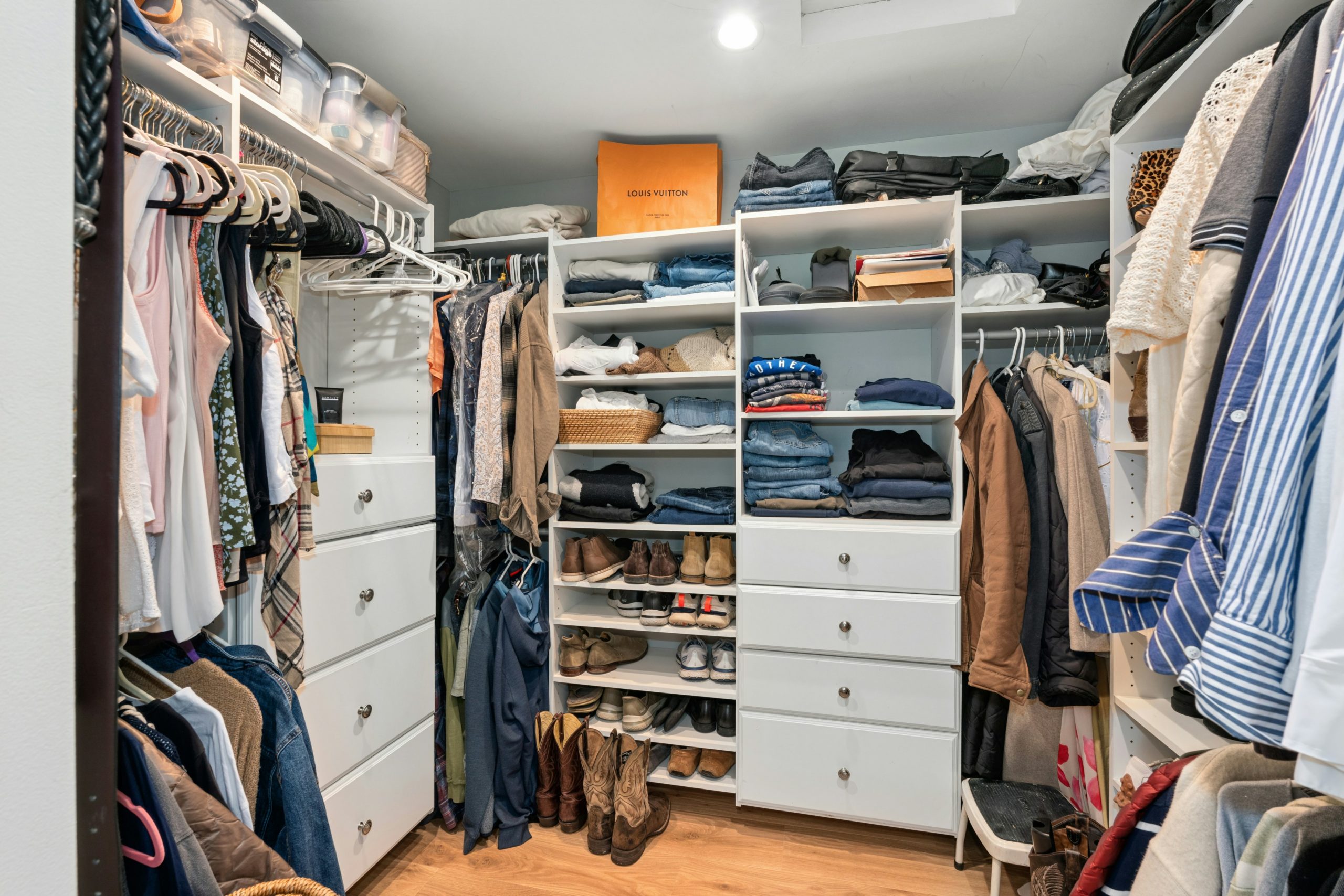 DSC08575 Walk-in closet with white built-in shelves, hanging clothes on both sides, folded jeans and shoes in the center, and an orange Louis Vuitton bag on the top shelf.